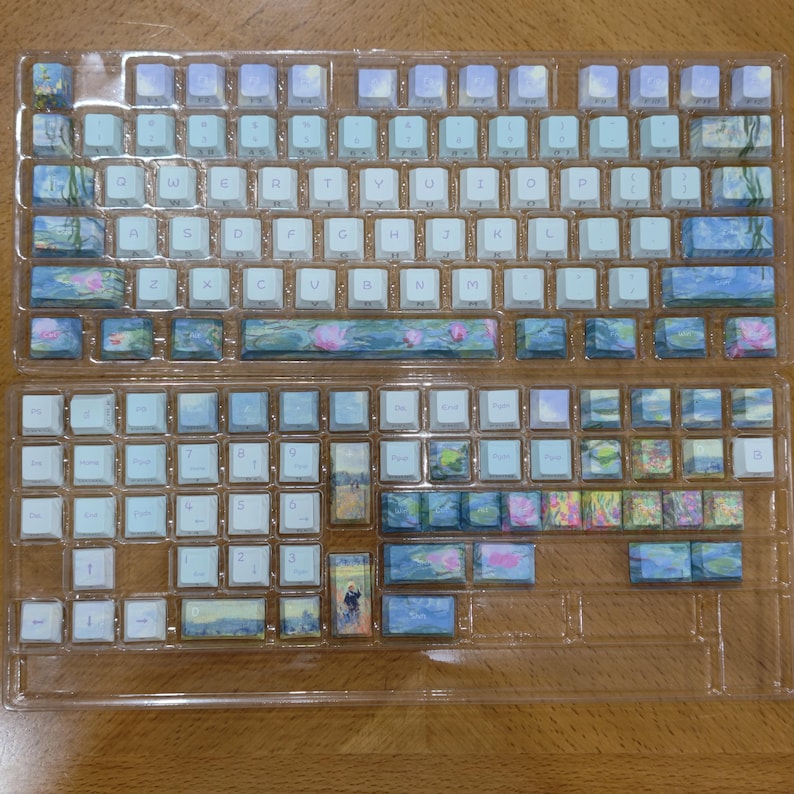 May include: A set of mechanical keyboard keycaps arranged in two clear plastic trays. The keycaps feature a white base with printed blue, green, and pink floral and landscape designs, including water lilies and figures in a field, with white letters.