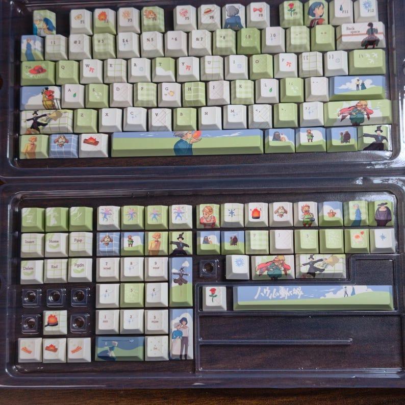 May include: A custom keyboard set with keycaps featuring anime-style illustrations and Japanese text. The keycaps are white and green, with various character designs. The keyboard is housed in a clear plastic case.