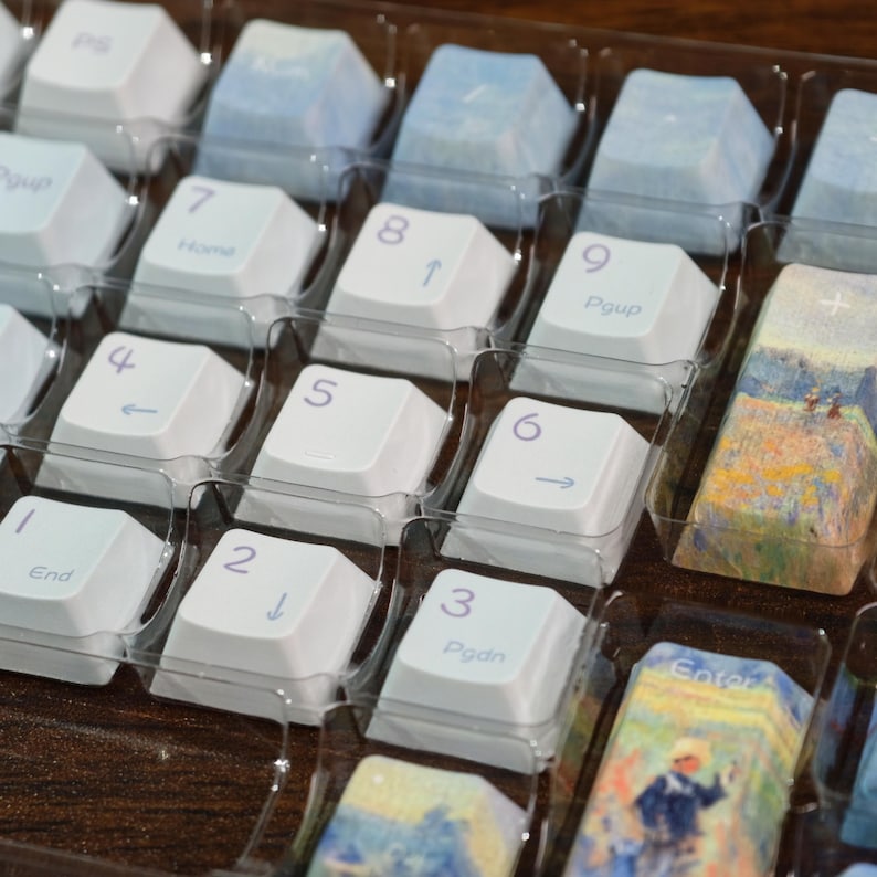 May include: A set of replacement mechanical keyboard keycaps arranged in a clear plastic tray. The keys include white numeric keypad caps with purple legends and several custom keycaps featuring colorful impressionist-style landscape paintings.