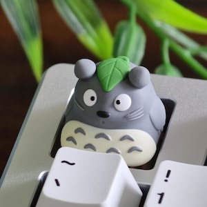 May include: A gray Totoro-shaped keycap with a green leaf on its head, set on a silver keyboard. The keycap has a white belly and black eyes. The keyboard keys are white.