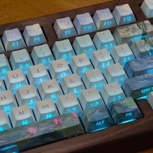 May include: A mechanical keyboard with a dark wood frame and white keycaps featuring blue backlit lettering. Several keys, including the spacebar, backspace, enter, shift, alt, and fn keys, display a colorful impressionist floral art pattern.