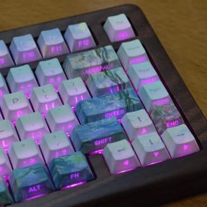 May include: A mechanical keyboard with a dark wood frame and white keycaps featuring pink backlighting. Several keys, including Backspace, Enter, Shift, and Fn, display a blue and green impressionist-style painting print with white text labels.