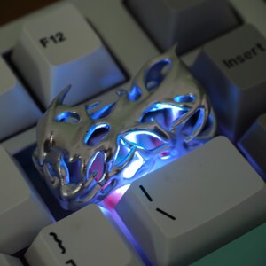 May include: A silver-colored, flame-shaped keycap with intricate cutouts, resting on a white keyboard. The keycap is illuminated with blue and pink light, creating a glowing effect. The keyboard keys are white with black lettering.