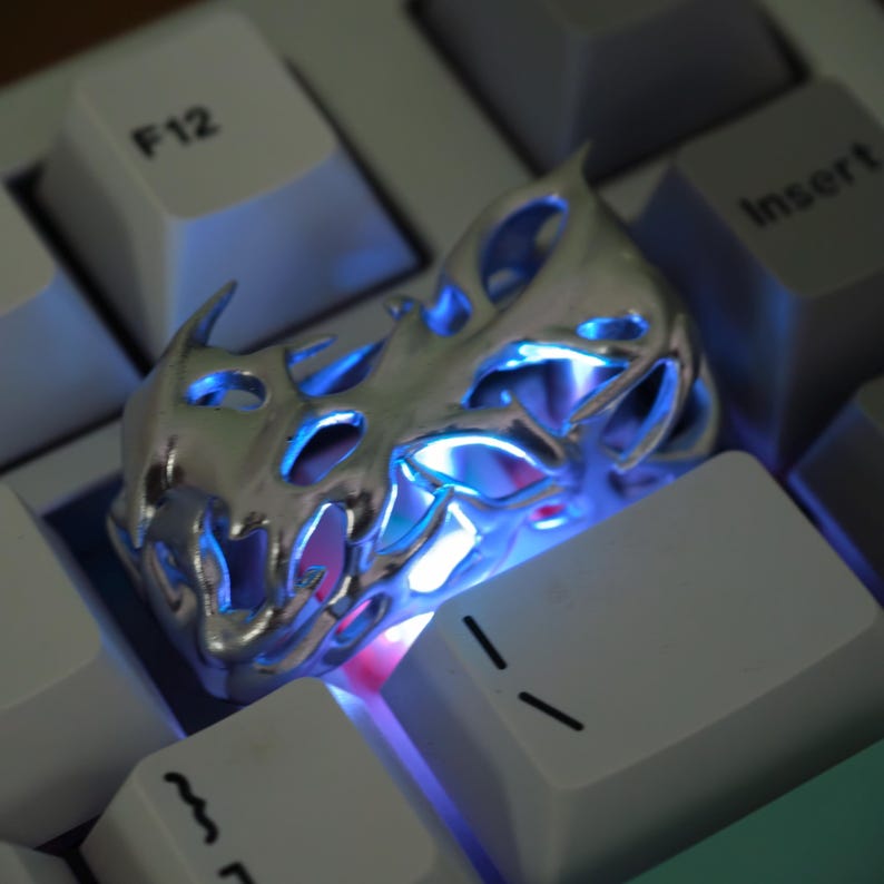 May include: A silver-colored, flame-shaped keycap with intricate cutouts, resting on a white keyboard. The keycap is illuminated with blue and pink light, creating a glowing effect. The keyboard keys are white with black lettering.