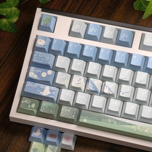 May include: A mechanical keyboard featuring a white frame and custom keycaps in shades of blue, green, and white. The keys display letters, numbers, and nature-themed illustrations including trees, sheep, flowers, and mountains, with some keys featuring cursive text.