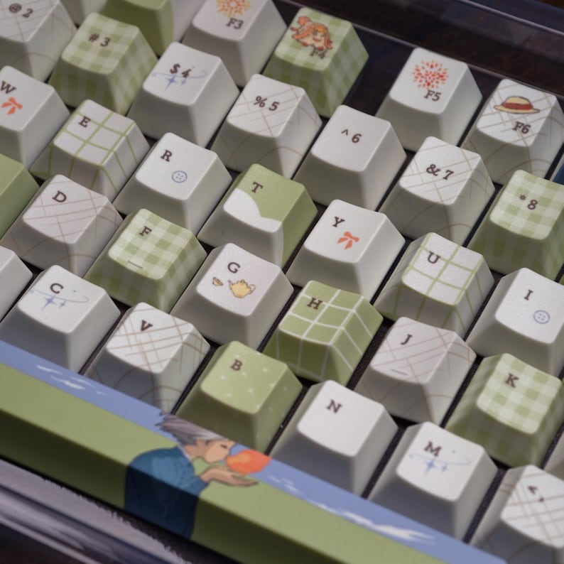 May include: Close-up of a custom keyboard with white and green keycaps. Some keycaps feature patterns like plaid and geometric designs, while others have small illustrations. The keyboard has a decorative strip with a painted scene.