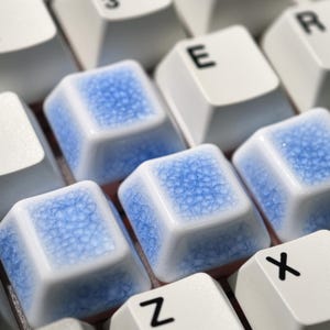 May include: Close-up of a computer keyboard with custom keycaps. The keycaps are white with a blue, crackled glaze design. The letters "E", "Z", "X", and "R" are visible on the keys.