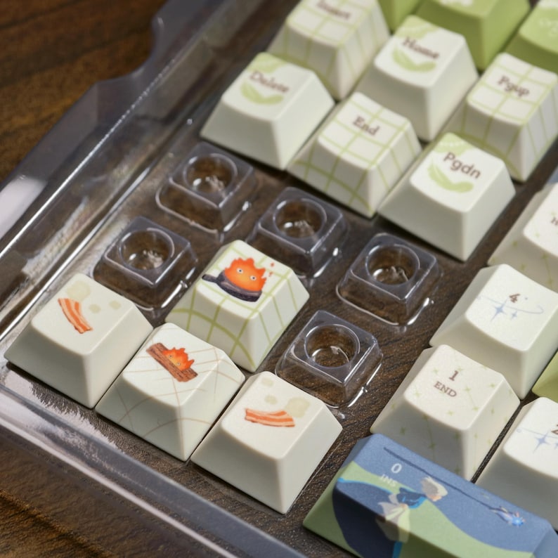 May include: A collection of custom keyboard keycaps in a clear plastic tray. The keycaps are white with various designs, including bacon, a campfire, and a cartoon character in a pan. Some keycaps have text like "End" and "Pgdn".