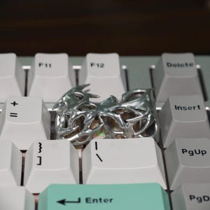 May include: A silver-colored ring with an intricate, organic design, featuring spiky protrusions and open spaces. The ring is displayed on a white keyboard, highlighting its unique texture and form.