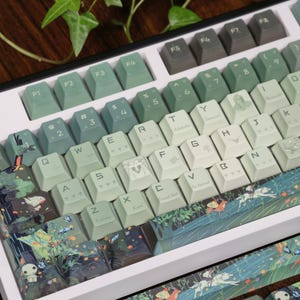 May include: A custom mechanical keyboard with green and white keycaps. The keycaps feature various designs, including forest scenes, butterflies, and character illustrations. The keyboard has a white frame and a variety of function keys.