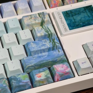 May include: A white mechanical keyboard featuring custom keycaps with impressionist-style floral and water lily paintings in shades of blue, green, and pink. The keyboard includes a small rectangular screen displaying the text SKYLOONG 30.