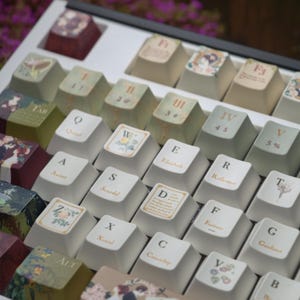 May include: A custom keyboard with keycaps featuring literary themes. The keycaps are off-white with various designs, including floral motifs, book titles, and quotes. The keyboard has a white frame and a selection of keycaps with different colors.