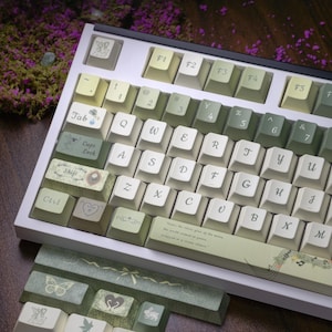 Może przedstawiać: Niestandardowa klawiatura mechaniczna z białą ramą i jasnozielonymi i kremowymi nakładkami na klawisze. Nakładki na klawisze mają dekoracyjne wzory i tekst, w tym cytat i nuty. Dodatkowe nakładki na klawisze są widoczne poniżej.