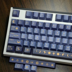 Pode incluir: Um teclado mecânico com moldura branca e teclas azul-escuras com ícones celestiais, numerais romanos e texto astronómico em dourado. A barra de espaço e teclas soltas adicionais exibem motivos de sol e lua num design dourado a condizer.