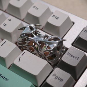 May include: A silver-colored ring with a unique, openwork design featuring sharp, pointed elements. The ring is placed on a computer keyboard with light gray keys, creating a contrast between the jewelry and the tech background.