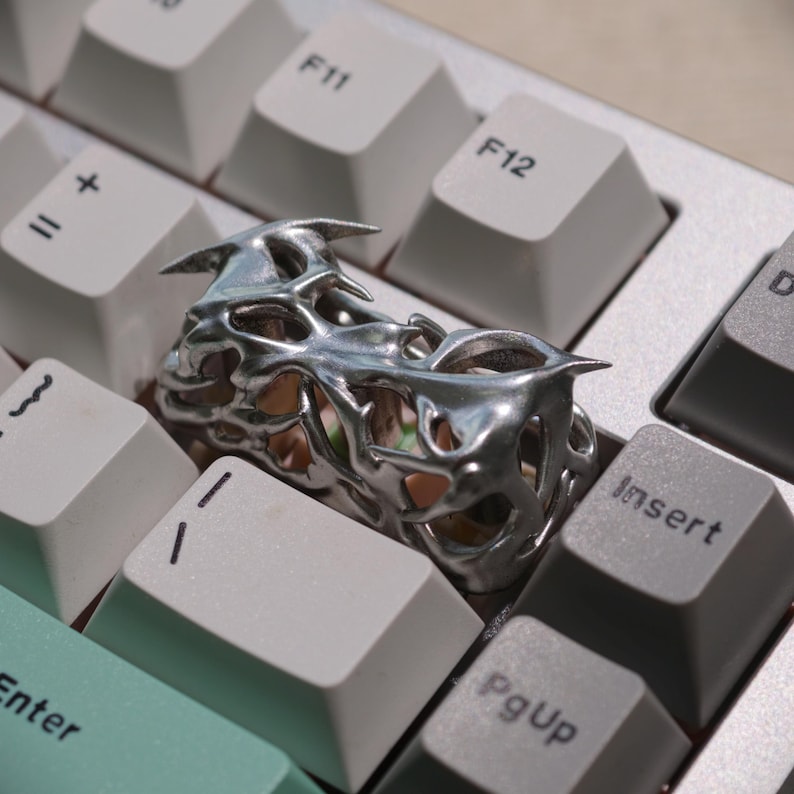 May include: A silver-colored ring with a unique, openwork design featuring sharp, pointed elements. The ring is placed on a computer keyboard with light gray keys, creating a contrast between the jewelry and the tech background.