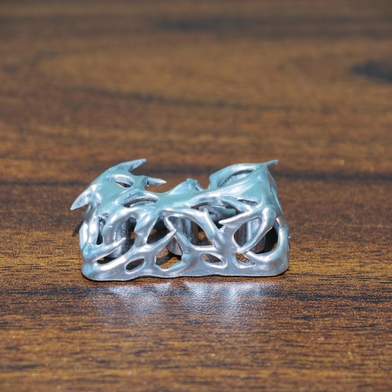 May include: A silver-colored ring with an intricate, openwork design. The ring features a series of interconnected, organic shapes, creating a textured and visually interesting surface. The ring is displayed on a wooden surface.