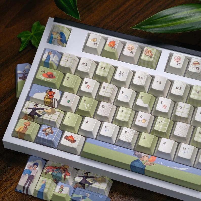 May include: A white mechanical keyboard with custom keycaps featuring various illustrations and patterns. The keycaps have a mix of colors, including green, white, and blue, with images of characters and objects. The keyboard is angled, with additional keycaps visible below.