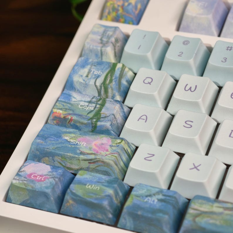 May include: A mechanical keyboard featuring a mix of solid light blue keycaps with purple lettering and custom keycaps decorated with an impressionist-style water lily painting. The keys include Tab, Caps, Shift, Ctrl, Win, Alt, Q, A, Z, 1, 2, 3, W, S, and X.