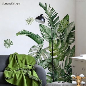 May include: A room with a tropical theme, featuring large green leaf wall decals and a black floor lamp. A gray sofa is adorned with a green blanket. The wall art includes large monstera leaves and other tropical foliage.
