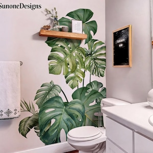 Puede incluir: Un baño con una gran calcomanía de pared de hojas de monstera verdes. Una estantería de madera contiene una pequeña imagen enmarcada y un jarrón de cristal con plantas. Inodoro y lavabo blancos visibles.