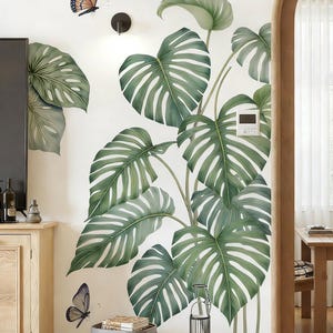Op de afbeelding: Een muursticker met grote groene monstera bladeren met witte nerven, wat een tropische uitstraling creëert. Twee vlinderstickers in oranje, blauw en wit zijn ook aanwezig. De tekst "SunoneDesigns" is zichtbaar.