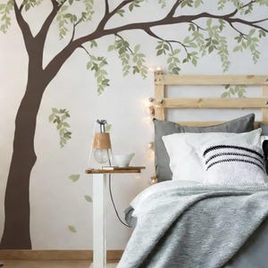 May include: A bedroom scene featuring a large tree wall decal in brown and green. A white nightstand with a lamp and a cup sits beside a bed with gray bedding and pillows. The headboard is made of light-colored wood.
