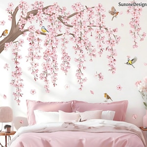 Op de afbeelding: Een roze slaapkamer met een muursticker van kersenbloesem. De sticker toont een bruine tak met roze bloemen en vogels. Het bed heeft roze beddengoed en witte kussens. De tekst "SunoneDesigns" staat rechtsboven.