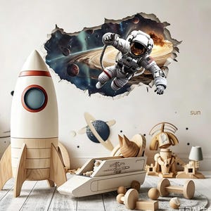 Può includere: Un adesivo da parete con un astronauta che fluttua nello spazio, un razzo giocattolo in legno, un veicolo spaziale giocattolo in legno e un robot giocattolo in legno. L'adesivo mostra un effetto 3D di un buco nel muro con una scena cosmica.