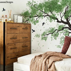 Könnte beinhalten: Eine Holzkommode mit sechs Schubladen, einem schwarzen Metallrahmen und dunklen Metallgriffen. Ein Wandbild eines Baumes mit grünen Blättern und schwarzen Vögeln ist zu sehen. Ein Bett mit einer braunen Decke und Kissen befindet sich im Vordergrund. Der Markenname "SunoneDesigns" ist sichtbar.