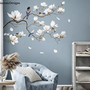 May include: Wall decal featuring a magnolia branch with white flowers and small birds. The decal is applied to a light blue wall, with a blue armchair and a side table in the foreground. The brand name "SunoneDesigns" is in the top left corner.