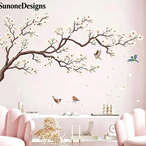 Op de afbeelding: Muursticker met een bloeiende tak met witte bloemen en bruine takken. Er worden verschillende vogels in vlucht afgebeeld, met vallende bloemblaadjes. De merknaam "SunoneDesigns" is linksboven zichtbaar.