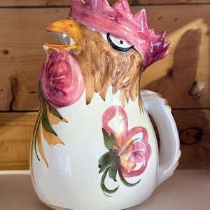 May include: A ceramic jug shaped like a rooster head with a pink comb, a yellow beak, and brown neck feathers. The body is white with hand-painted pink and green floral motifs. It features a curved handle on the side and a wide spout.