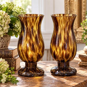 May include: A pair of matching glass vases featuring a flared rim, a bulbous body, and a circular pedestal base. The glass has a translucent amber and dark brown mottled pattern that resembles tortoiseshell, creating a distinct visual texture.