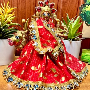 May include: A gold-colored statue of a deity, adorned with a red and gold sari. The statue has multiple arms and is seated on a golden lion. The sari is embellished with gold and blue accents. The deity wears a jeweled crown.