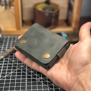 Op de afbeelding: Een compacte olijfgroene lederen portemonnee met twee messingkleurige metalen drukknopen op de voorflap. De portemonnee heeft een gladde, matte afwerking en een rechthoekige vorm met afgeronde hoeken, ontworpen om in de handpalm te passen.