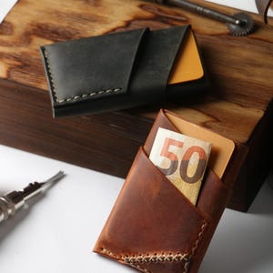 Könnte beinhalten: Zwei Leder-Kartenetuis. Eines ist dunkelgrau, das andere braun. Im braunen Kartenetui ist ein Fünfzig-Euro-Schein teilweise sichtbar. Beide haben sichtbare Nähte und ein minimalistisches Design.