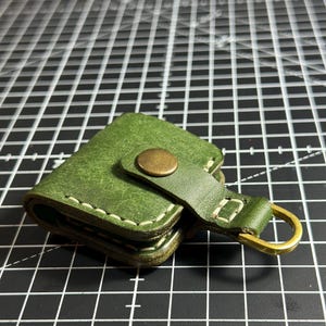 Könnte beinhalten: Ein kleines, quadratisches Etui aus olivgrünem Leder mit sichtbaren cremefarbenen Nähten an den Kanten. Das Etui wird mit einem messingfarbenen Metalldruckknopf an einem Lederriemen verschlossen, der einen messingfarbenen D-Ring enthält.
