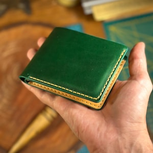 Könnte beinhalten: Ein Faltgeldbörse aus glattem, dunkelgrünem Leder mit kontrastierenden hellbraunen Kanten und sichtbaren, dicken cremefarbenen Nähten entlang des Randes. Die Geldbörse hat eine klare, rechteckige Form mit abgerundeten Ecken.