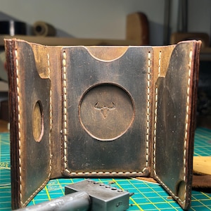 May include: A tri-fold brown leather wallet featuring visible light-coloured stitching, three card slots with rounded cutouts, and a central circular embossed bull skull logo. A metal leathercraft tool with the number 5 is positioned in the foreground.