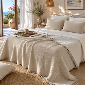 May include: A beige textured bedspread with fringe trim draped over a bed featuring two matching rectangular pillows, a white flat sheet, and a white duvet. The bedding set is arranged with a small wooden tray and an open book on top.