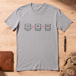 May include: A light gray short-sleeved t-shirt featuring a graphic of three computer keyboard keys labeled ctrl, alt, and del. Below the keys, the words focus, mindeet, and excuees are printed in black. Red plus signs separate the keys.