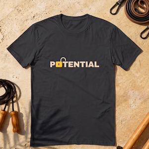 May include: A dark gray short-sleeve t-shirt featuring the word POTENTIAL printed in light beige letters across the chest. The letter O is replaced by a yellow padlock icon with an open shackle, creating the visual wordplay of unlocked potential.