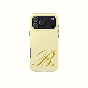 Puede incluir: Funda de teléfono amarillo pálido con la letra "B" dorada en escritura cursiva. La funda tiene un módulo de cámara negro con tres lentes y un flash. La funda es de color liso con un diseño sencillo y elegante.