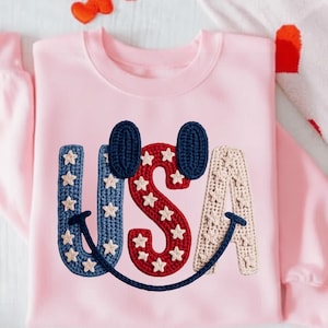 Può includere: Una felpa rosa chiaro con la scritta "USA" in un design all'uncinetto. La "U" e la "A" sono blu e beige, rispettivamente, con stelle bianche. La "S" è rossa con stelle bianche. Il design include due orecchie all'uncinetto blu navy e un sorriso nero.