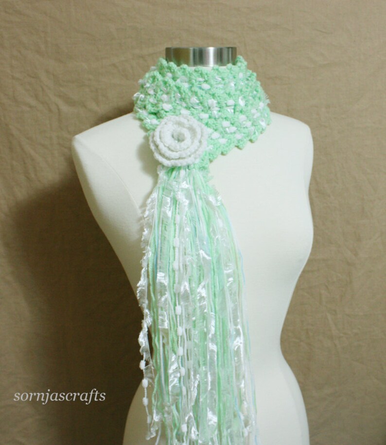 May include: A green and white crocheted scarf with a white crocheted flower accent. The scarf is made of a soft, lightweight yarn and has a fringed edge.