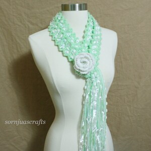 May include: A light green crocheted scarf with a white crocheted flower accent. The scarf has a long, flowing fringe.