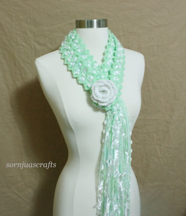 May include: A light green crocheted scarf with a white crocheted flower accent. The scarf has a long, flowing fringe.