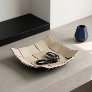 May include: A beige and black woven leather tray holds a car key with a black leather keychain. A small blue vase is nearby. The square tray has a woven design, ideal for storing small items.