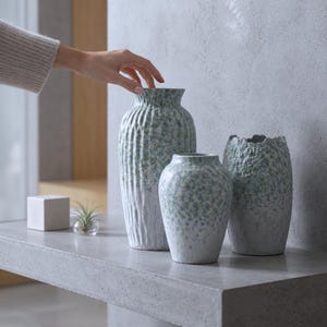 May include: Three ceramic vases with a textured, white and sage green speckled finish. The tallest vase features vertical ridges, while the two shorter vases have a rough, organic surface texture and rounded shapes with uneven, jagged rims.